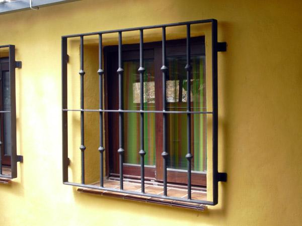 Finestra con grate nere su muro giallo, vetri riflettenti e luci naturali che illuminano l’ingresso esterno.