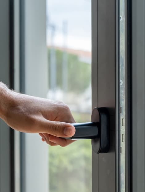 Mano che apre una finestra a scorrimento, luce naturale entra, cornice metallica scura e vetro riflettente.